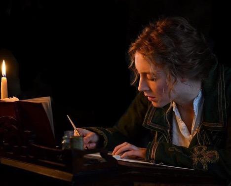 In a still shot from Little Women, Saoirse Ronan as Jo, sits by candlelight writing at a desk